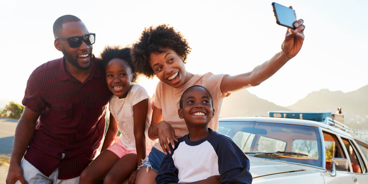 African American family posing for selfie next to car packed for road trip with car insurance