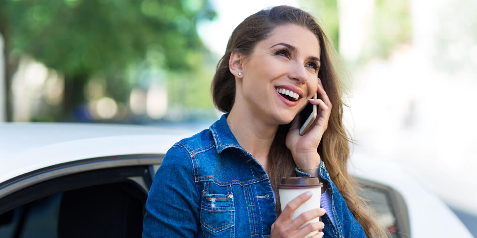 smiling woman holing a coffee leaning on a car talking on smartphone