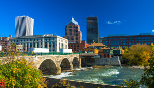 Downtown Rochester skyline in the background with a stone bridge, the Genesee River, and plants in the foreground.