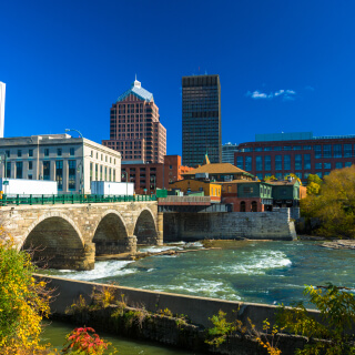 Downtown Rochester skyline in the background with a stone bridge, the Genesee River, and plants in the foreground.