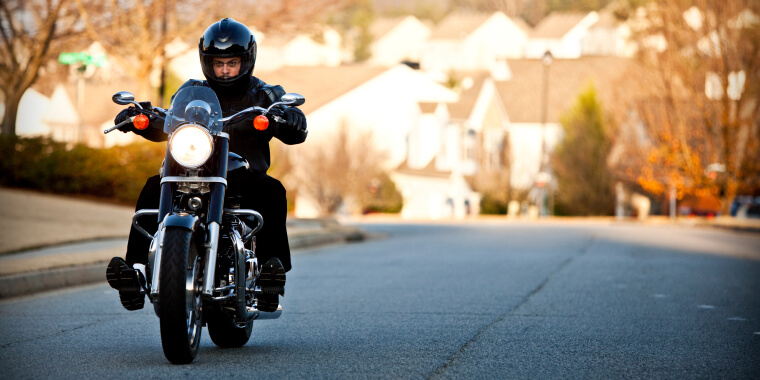 A man rides his insurance-covered motorcycle in full protective gear on a quiet residential street.