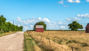 An abandoned old barn in Rural Texas with the state flag painted on its roof sits in a farmland community framed by fields and a dirt road: Levelland cheap car insurance in The Lone Star State.