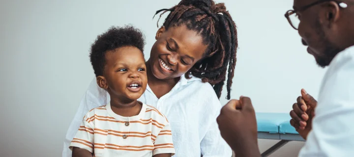 A joyful black boy sits on his mother's lap during a medical consultation as the doctor engages them in conversation, ensuring a soothing healthcare environment.