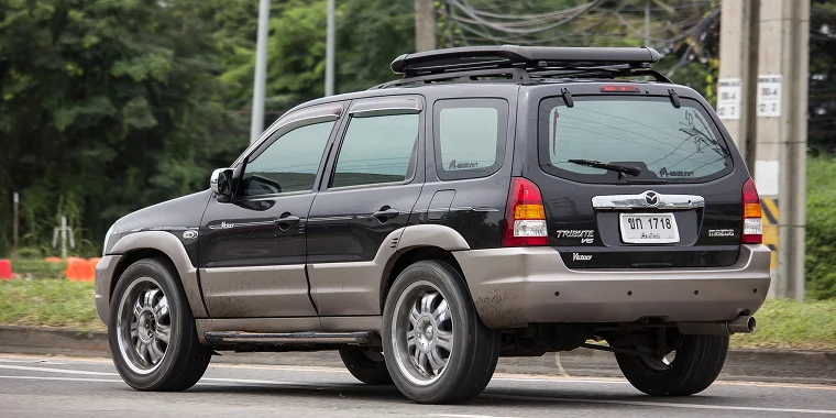 Mazda Tribute black with gray advancing in front of a road with trees on a cloudy day in Chiangmai, Thailand – Mazda, cheap car insurance.