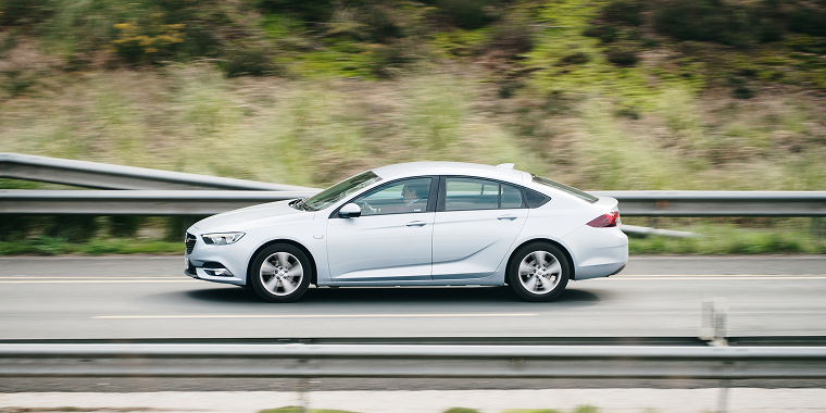 White Saturn Aura driving on a road at speed, with a wooded background during the day. Cheap car insurance.