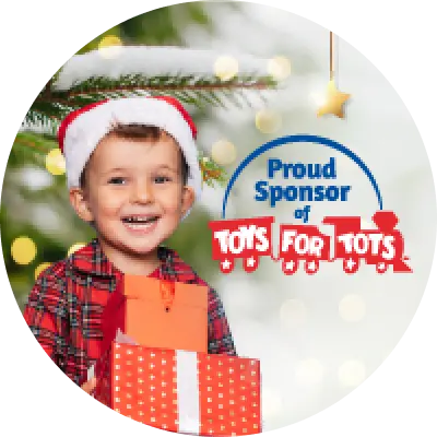A smiling child in a red plaid shirt and Santa hat holds wrapped presents in front of a snowy Christmas tree, next to text that reads “Proud Sponsor of Toys for Tots.”