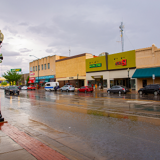 Rainy street scene in downtown Roswell, New Mexico, with parked cars, storefronts, wet sidewalks, and reflections on the road after a passing shower.