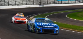 Two NASCAR race cars speed around a track, including a blue Freeway Insurance branded car in the lead.
