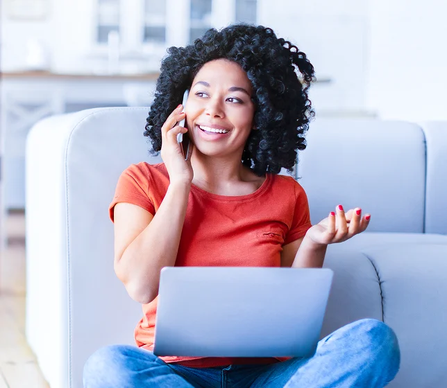 A smiling woman sits on a couch using a laptop while talking on her phone, illustrating smart side-by-side comparisons and saving time.