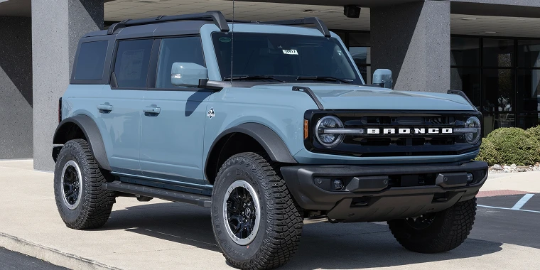 Light blue ford bronco SUV with off-road tires and a black hardtop parked on a concrete curb in front of a modern building — Cheap car insurance.
