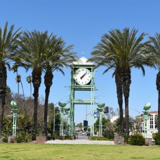 Clock tower surrounded by palm trees in Village Green Park, Garden Grove, California, under a clear blue sky. Cars and low-rise buildings line the street in the background.
