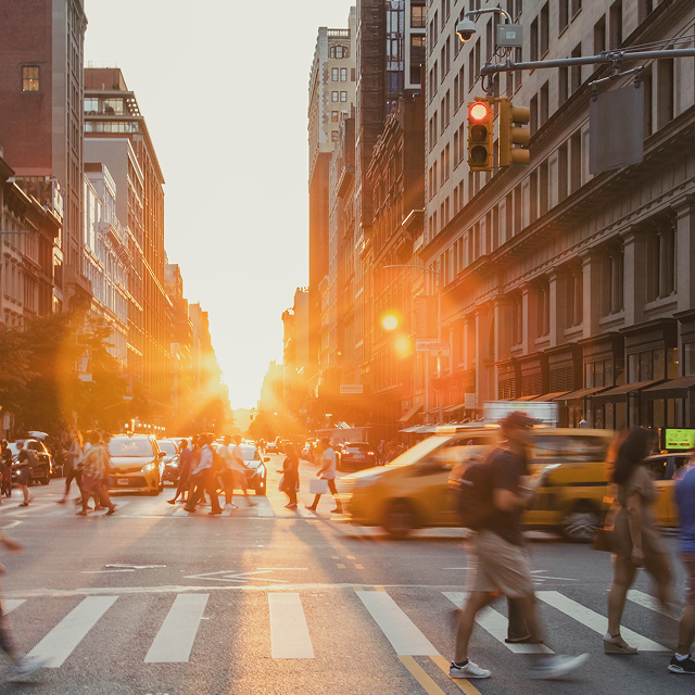 Alt Text A busy intersection on 5th Avenue and 23rd Street in New York City is crowded with people and cars, with a sunset background.