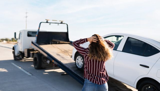 Woman in a striped sweater stands with hands on her head as a tow truck loads her white car onto the flatbed, showing what to do to prevent car from being towed after a roadside breakdown.