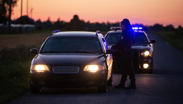 Police officer speaking to a driver during a traffic stop at sunset, requesting proof of car insurance.