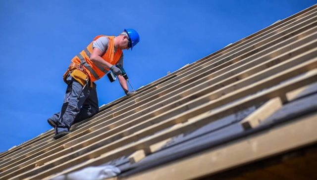 Worker installing wooden roof framing during roof replacement under clear blue sky.