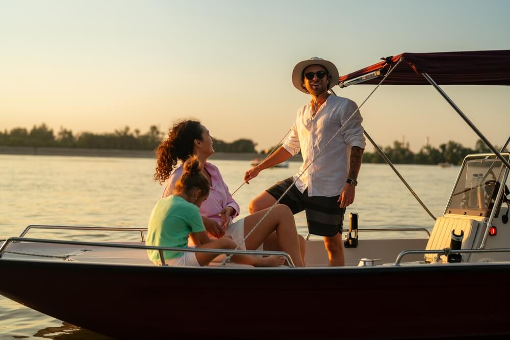 A family enjoying a boat ride with the best boat insurance near me on a calm lake during sunset. The man is standing and smiling, while the woman and child are seated and looking at him.