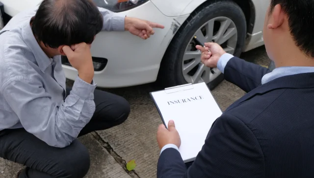 Insurance adjuster in a suit reviews an insurance clipboard and points to scuff marks on a car bumper while the worried driver crouches nearby, showing how to file a car insurance claim.