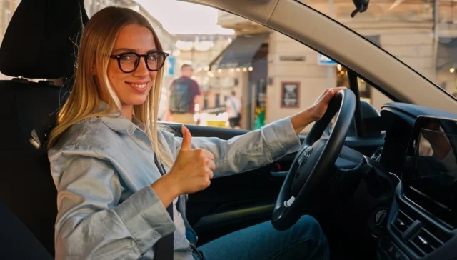 Smiling woman with glasses sits in the driver’s seat and gives a thumbs-up, suggesting smart choices that can lower car insurance premium.