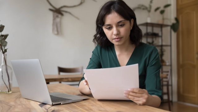 Woman at home reading a bill beside her laptop while researching options to compare car insurance quotes.