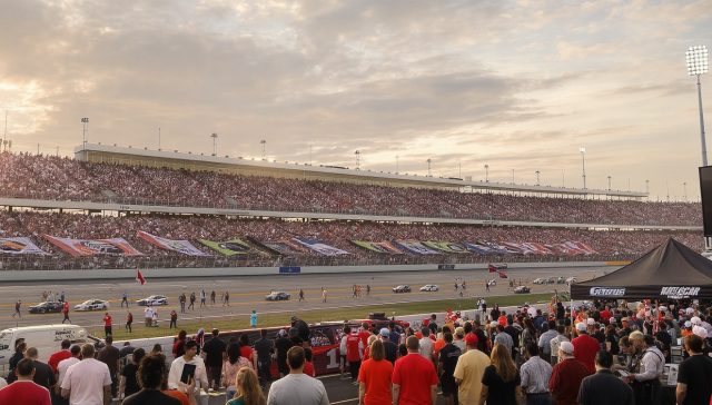 A vast nascar infield area under an autumnal sky, vibrant with energy and laughter. Below fluttering nascar team banners, hundreds of adults from 30-60 years old, clad in nascar colors buzz like a playful hive