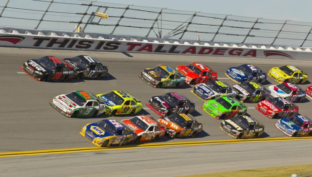 NASCARs racing in a tight pack at Talladega Superspeedway, with colorful stock cars drafting side-by-side on the high-banked track and “This Is Talladega” visible on the wall in the background.