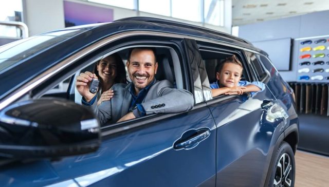 A joyful family sits in their new car at a dealership. The father holds the key, while the mother and child share the excitement of a new vehicle acquisition since their car insurance rates dropped.