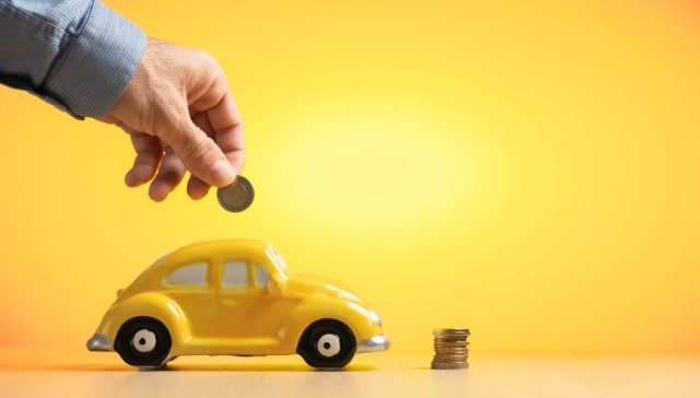 Closeup hand putting coin into a miniature yellow car piggy bank, saving on car insurance, saving workshop expenses, saving fuel, broken down car, selling or renting a car, free background yellow