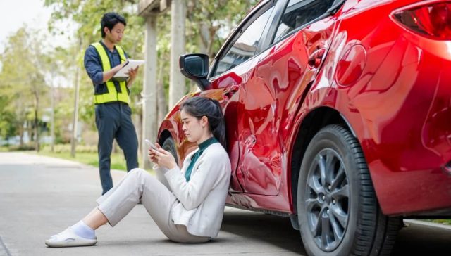 Woman sitting beside a red car with major side damage after an accident while an insurance adjuster documents the scene, illustrating a car insurance record claim.