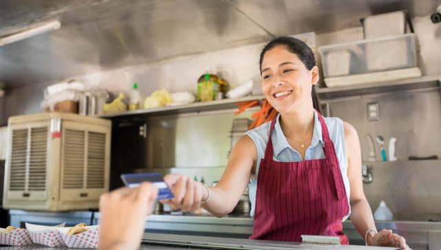 A smiling food truck employee in a red apron hands a customer a credit card payment through the service window inside a stainless-steel food truck kitchen
