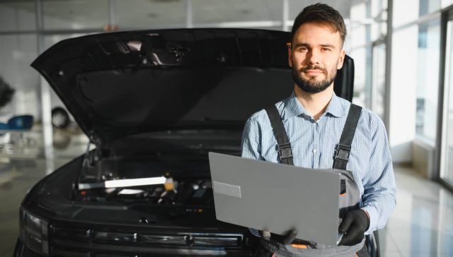 Certified technician using a laptop to diagnose vehicle software repairs with the hood open in a professional service center.