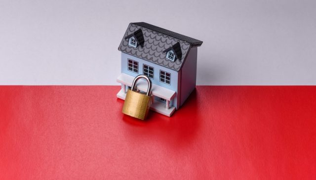 Miniature model house secured with a brass padlock on a red and white background, representing smart home insurance, home security, and property protection.