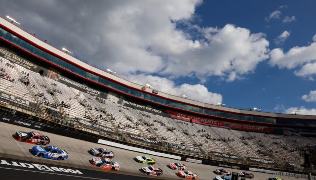 Bristol NASCAR race with a pack of stock cars speeding around the steeply banked track, grandstands in the background, and a bright blue sky filled with large clouds.