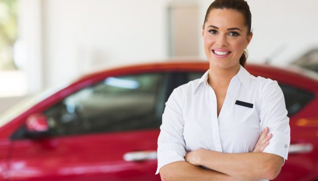 Smiling automotive professional standing beside a red car for a Compliant Driver Program.