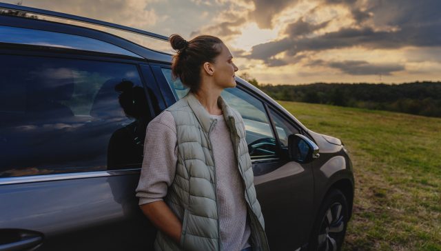 Young female driver standing by car enjoying sunset and nature in field during road trip.