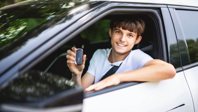 Smiling young driver holding a car key while sitting behind the wheel of a white vehicle, illustrating new driver coverage costs.