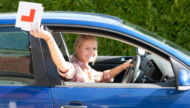 Young driver holding a learner’s permit sign while sitting behind the wheel of a car.