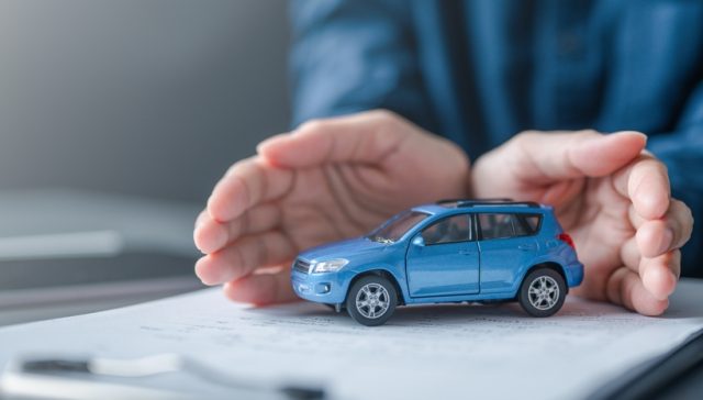 Hands protecting a blue model car on a desk, representing car insurance reinstatement and coverage protection.